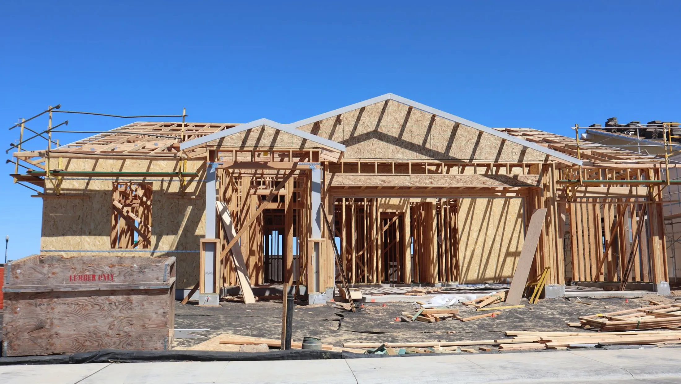 A single-storey house under construction, framed in dimensional lumber with roof trusses set and gable sheathing fixed, under a clear sky